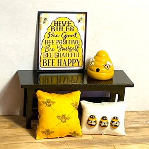 May include: A black framed sign with the text "Hive Rules Bee Good Bee Positive Bee Yourself Bee Grateful Bee Happy" in yellow and black letters on a yellow and black striped background. A yellow ceramic beehive with bees is on a black table with two pillows, one yellow with bees and one white with bees.