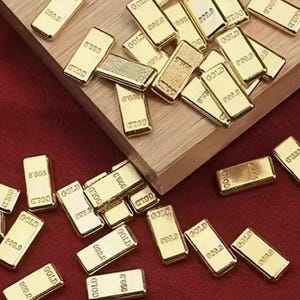 May include: A collection of small, rectangular gold bars scattered on a wooden surface and a red fabric. Each bar is stamped with the word "GOLD" and a purity marking of "999.9".