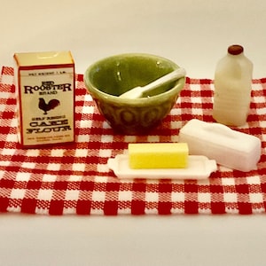 May include: A miniature kitchen set featuring a box of Red Rooster Brand self-rising cake flour, a green mixing bowl with a white spoon, a white milk jug, and a yellow stick of butter on a white dish, all arranged on a red and white checkered cloth.