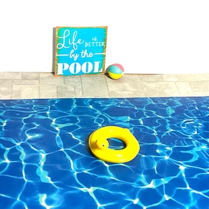 May include: A blue pool with a yellow inflatable ring and a multi-colored beach ball. A wooden sign with the text "Life is better by the pool" is on the wall behind the pool.