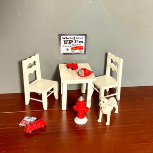 May include: A white table and two chairs with a red fire truck, fire hydrant, and a Dalmatian dog figurine. A red fire hat and a sign that reads "When I grow UP I'm going to drive a fire truck" are on the table.