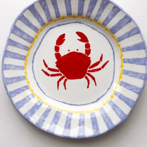 Könnte beinhalten: Ein weißer Keramikteller mit blauen und gelben Streifen. Ein roter Krebs ist in die Mitte des Tellers gemalt.