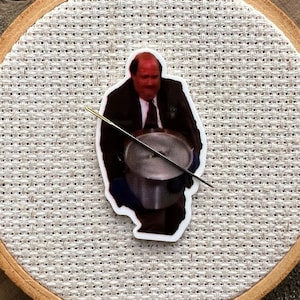 May include: A round wooden embroidery hoop with a white fabric background. A sticker of a man in a suit holding a pot is centered on the fabric.