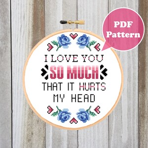 Puede incluir: Un patrón de punto de cruz con un aro blanco con el texto "I love you so much that it hurts my head" en letras rosa y azul. El aro está decorado con rosas azules y corazones rosas.