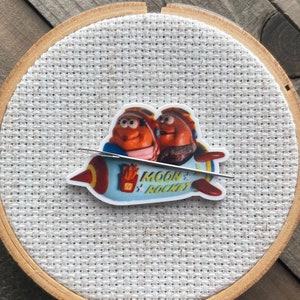 Pode incluir: Um bastidor de bordado de madeira com uma imagem de desenho animado de dois personagens em um foguete. O foguete é azul e vermelho com as palavras "Moon's Rocket". O fundo é de tecido branco.