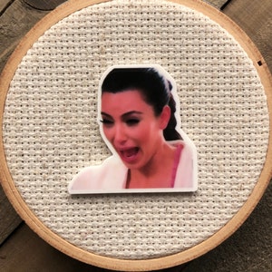 Puede incluir: Un aro de bordado blanco con un fondo de tela beige. Una imagen impresa de una mujer con cabello oscuro y una expresión de sorpresa está centrada en el aro.
