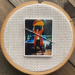 May include: A white embroidery hoop with a brown wooden frame. Inside the hoop is a printed image of a man holding a large burrito above his head. The man is wearing a brown coat and jeans. The image is on a white background.