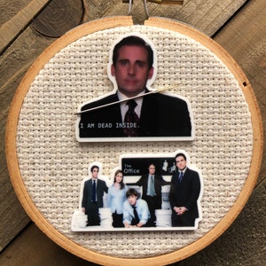 May include: A round wooden embroidery hoop with white fabric stretched across the center. The fabric features two images from the TV show "The Office". The top image is a close-up of the character Michael Scott with the text "I am dead inside." The bottom image shows a group of characters from the show standing in front of a sign that reads "The Office".
