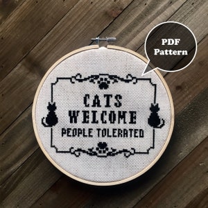 Puede incluir: Un aro de bordado blanco con un diseño de punto de cruz en blanco y negro que dice "Cats Welcome People Tolerated". El diseño incluye dos gatos negros y una huella de pata.