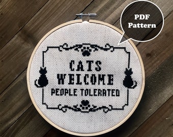 May include: A white embroidery hoop with a black and white cross-stitch design that reads "Cats Welcome People Tolerated". The design includes two black cats and a paw print.