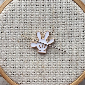 May include: A white and gold needle minder shaped like a cartoon hand with four fingers. The needle minder is on a beige fabric background.