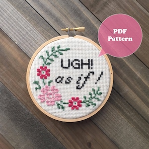 May include: A white embroidery hoop with a cross-stitch design featuring the phrase "UGH! as if" surrounded by green leaves and pink and red flowers. The hoop is made of wood and has a gold metal screw.