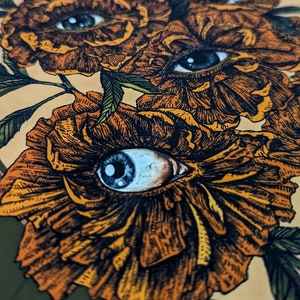 Photo of an art print showing hand drawn golden flowers with photorealistic eyes at the centre of each.