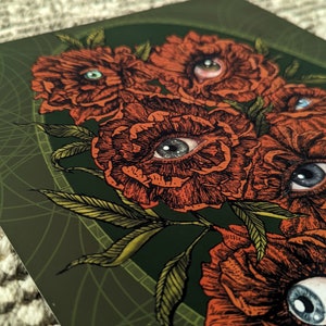 close up photo of an art print of a hand drawn bouquet of scarlet flowers with photorealistic eyes at the centre of each with a deep green background.  surrounded by an olive green oval frame  with leaf details.