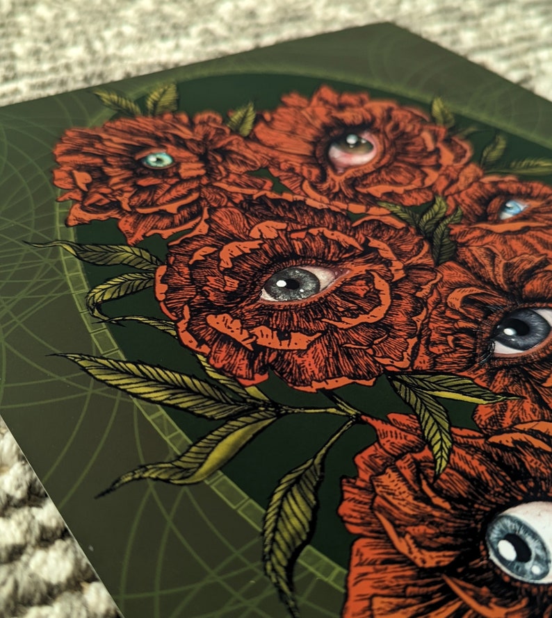 close up photo of an art print of a hand drawn bouquet of scarlet flowers with photorealistic eyes at the centre of each with a deep green background.  surrounded by an olive green oval frame  with leaf details.