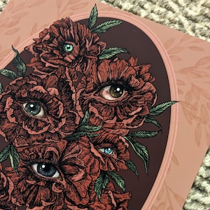 close up photo of an art print of a hand drawn bouquet of pink red  flowers with photorealistic eyes at the centre of each with a burgundy background.  surrounded by mauve pink oval frame  with leaf details.