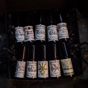 May include: A set of ten small glass bottles with black roll-on tops. Each bottle has a white label with black text. The labels feature names like "The Depth", "The Stad", "Coin-Opera Boy", "The Moon", "Uranus", "The Hermit", "Drackus", "Vega, Deneb & Altair", "Blood Moon", and "The Impero".