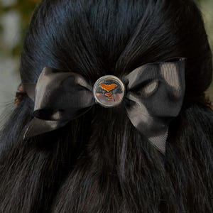 May include: A dark grey hair bow made of ribbed fabric featuring a circular silver-toned metal centre piece. The centre piece displays a small orange bat graphic with a subtle gradient effect, centred on the bow to secure the fabric loops.