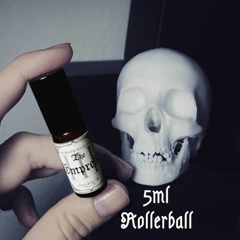 Puede incluir: Un frasco de 5 ml de aceite de perfume en bola con una etiqueta negra que dice "Andromeda's Garden The Empress II Perfume Oil". El frasco se sostiene en una mano contra un fondo de calavera blanca.