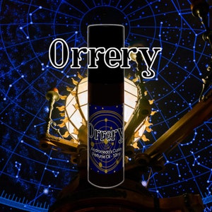 Op de afbeelding: Een donkerbruine glazen fles met een zwarte dop en een blauw en gouden etiket. Het etiket vermeldt "Orrey Andromeda's Curse Perfume Oil - 10ml". De fles staat voor een donkerblauwe achtergrond met witte sterren.