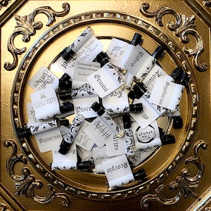 May include: A collection of small glass perfume oil sample vials with black caps, each featuring a white label with black text including names like Gemini, Scorpio, and Aquarius, arranged in a circular gold-toned decorative frame.