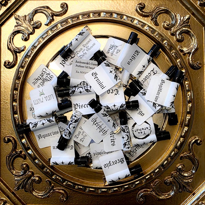 May include: A collection of small glass perfume oil sample vials with black caps, each featuring a white label with black text including names like Gemini, Scorpio, and Aquarius, arranged in a circular gold-toned decorative frame.