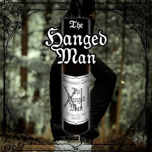 Könnte beinhalten: Eine schwarze Parfümflasche mit dem Text "The Hanged Man" in weißer Schrift. Die Flasche hat ein silbernes Etikett mit "Andromeda's Curse" und "Perfume Oil 10ml". Der Hintergrund ist eine verschwommene Waldszene.