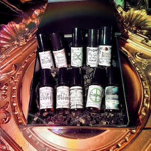 May include: A selection of small perfume bottles with black lids and white labels, presented in a decorative tray. The labels bear names such as "The Devil," "Briar Rose," and "Earth." The bottles are set on a bed of dark, shredded material.