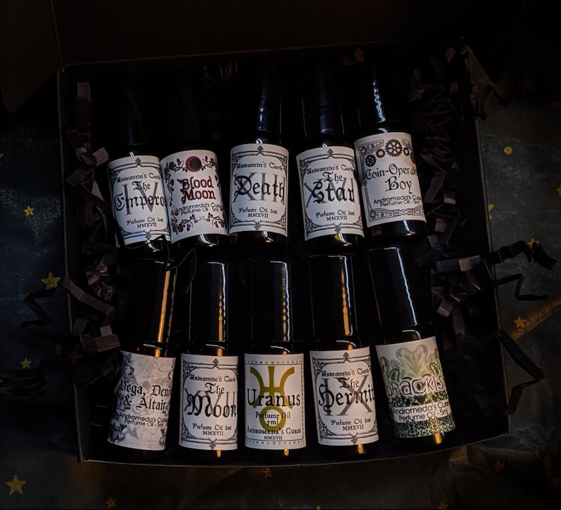 May include: A set of ten small glass bottles with black roll-on tops. Each bottle has a white label with black text. The labels feature names like "The Depth", "The Stad", "Coin-Opera Boy", "The Moon", "Uranus", "The Hermit", "Drackus", "Vega, Deneb & Altair", "Blood Moon", and "The Impero".