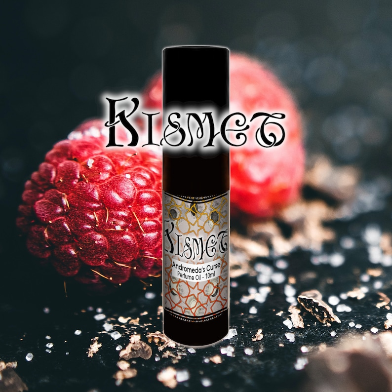 May include: A black roller bottle with a gold label that reads "Kismet Andromeda's Curse Perfume Oil - 10ml". The bottle is sitting on a dark surface with a red raspberry in the background.