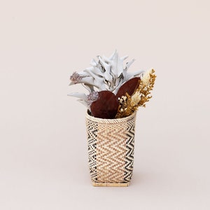 May include: A woven basket with a geometric pattern holds a bouquet of dried flowers. The flowers are white, brown, and beige.