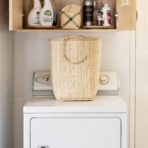 May include: A white washing machine with a natural woven basket on top. The washing machine has a white door with a handle. The basket is made of natural fibers and has a woven pattern. The basket is sitting on top of the washing machine.