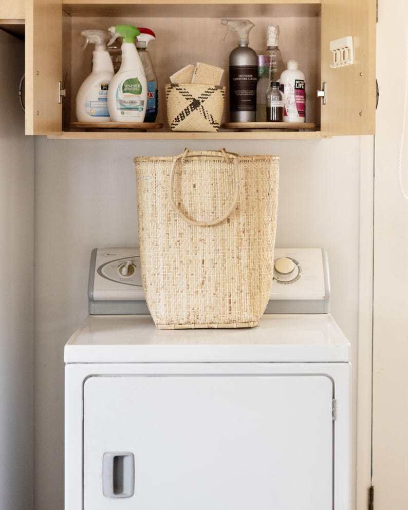 May include: A white washing machine with a natural woven basket on top. The washing machine has a white door with a handle. The basket is made of natural fibers and has a woven pattern. The basket is sitting on top of the washing machine.