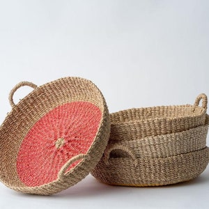 May include: Woven oval baskets with handles. One basket features a coral-colored center. The baskets are stacked, showcasing their natural fiber construction and design. Ideal for storage or decor.