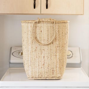 May include: A large, natural woven basket with a single, thick, rounded handle. The basket is made of light brown, natural fibers and is sitting on a white surface.