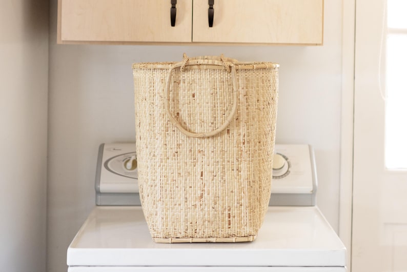 May include: A large, natural woven basket with a single, thick, rounded handle. The basket is made of light brown, natural fibers and is sitting on a white surface.