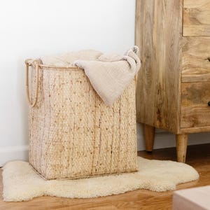 May include: A woven wicker basket with a natural finish, filled with a light beige blanket. The basket has two handles and is sitting on a white fluffy rug.
