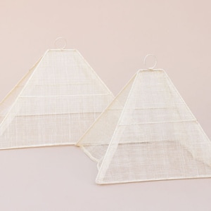 May include: Two white, pyramid-shaped food covers made of mesh fabric. The covers are made of a fine, woven material and have a circular metal ring at the top.