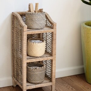 May include: A three-tiered woven rattan shelf unit with three woven baskets. The top basket has a black and white geometric pattern, the middle basket is natural woven, and the bottom basket has a black and white striped pattern. The shelf unit is standing on a wooden floor.