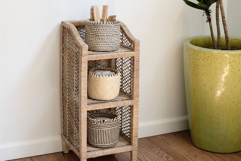 May include: A three-tiered woven rattan shelf unit with three woven baskets. The top basket has a black and white geometric pattern, the middle basket is natural woven, and the bottom basket has a black and white striped pattern. The shelf unit is standing on a wooden floor.