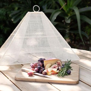 Op de afbeelding: Een witte, piramidevormige voedselafdekking met een ringhandvat staat op een houten tafel. Een houten snijplank bevat druiven, kaas, gesneden appels, vijgen en rozemarijn. Een mes ligt op de plank. De scène is buiten met groen op de achtergrond.