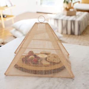 May include: A beige mesh food cover in a pyramid shape, protecting a wooden serving board with fruit and crackers. The cover has a metal ring on top for easy handling. The board sits on a white marble surface.