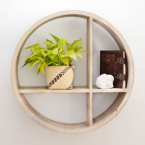 May include: A round, light-colored wooden shelf with four compartments. The shelf holds a potted plant with bright green leaves, a brown leather-bound journal, and a white rock. The shelf is mounted on a white wall.