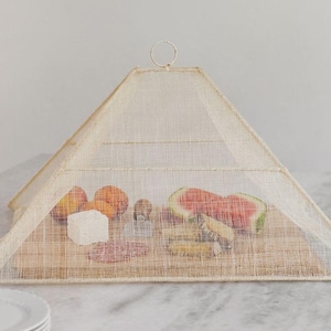 Könnte beinhalten: Ein beiger, pyramidenförmiger Lebensmittel-Schutz aus gewebtem Material, der eine Präsentation von Obst, Käse und Charcuterie schützt. Der Deckel hat einen kleinen Metallring an der Spitze. Geschnittene Wassermelone, Pfirsiche und andere Snacks sind sichtbar.