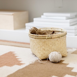 May include: A woven basket with a natural finish sits on a patterned rug. The basket is filled with twine and a ball of yarn.