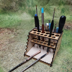 May include: Wooden paintbrush holder with multiple slots for storing brushes. The holder has a tiered design with two levels. Several paintbrushes are in the holder, including a blue brush, a black brush, and a silver brush.