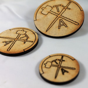 May include: Three wooden circles with a black border and a laser-etched design of a hammer and two swords crossed with the letters "TTA" in the center.