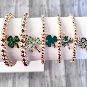 May include: Five gold bracelets with green shamrock charms. The bracelets are made of small gold beads and are layered on a white surface.
