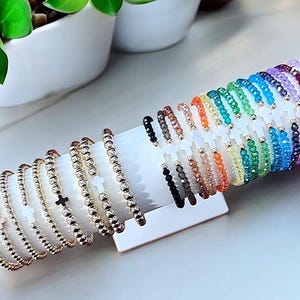 May include: A display of colorful beaded bracelets with a white cross charm. The bracelets are arranged in a rainbow pattern on a white stand.