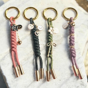 May include: Four keychains with gold-colored rings and accents. Each keychain features a different color of braided cord: pink, black, green, and purple. Charms include flowers, smiley faces, hearts, and initial letters. The keychains have gold-colored cylindrical end caps.